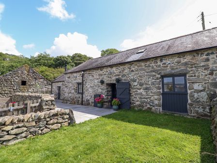 A stone cottage with slate roof and black doors and windows with potted plants on a paved area and a stone wall in the garden at Beudy Bach in Llanfair near Harlech