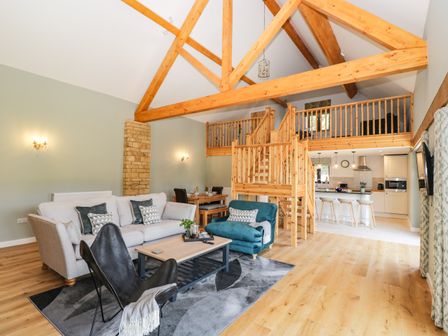 An open living space with wooden beams a staircase leading to a loft kitchen with bar stools and a seating area with sofas and a lounge chair at Petals Patch in Lower Slaughter