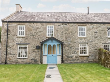 A stone house with a blue door and multiple windows with a grassy front yard and a paved pathway leading to the entrance at 8 Llwyn Onn in Llanfairpwllgwyngyll