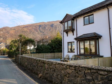 A white house with brown framed windows behind a stone wall next to a road with mountains in the background at Aran in Beddgelert