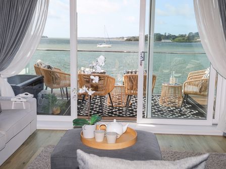 A living room with a teapot and cups on a tray on an ottoman facing a balcony with wicker chairs and a view of water at Seaclusion in Y Felinheli
