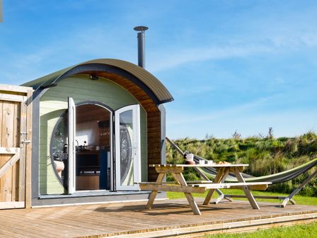A wooden cabin with curved roof and open doors next to a picnic table and hammock on a wooden deck at Dew Pond in Blagdon
