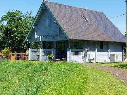 A small wooden house with a steep roof and a covered porch surrounded by grass and trees at Orchard Lodge in Hockworthy near Sampford Peverell Devon