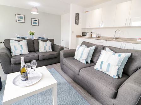 A living area with two gray sofas with pillows a white table with a bottle and glasses and a kitchen area in the background at Apartment 6 in Llandudno