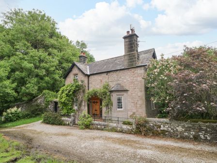 A stone house with two chimneys surrounded by trees and plants at The Lodge in Greystoke