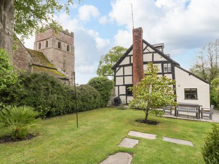 A garden with grass a tree stone path and a patio area next to a Tudor style house and a stone church tower in the background at Notts House in Wellington
