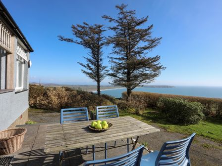 A patio with a wooden table and blue chairs overlooking trees and the sea at Bryn Meillion in Rhiw near Sarn Meyllteyrn