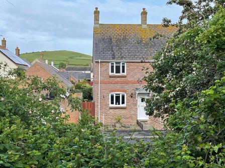 A brick house with white windows and door surrounded by green bushes and trees with a hill in the background at The Beach House in West Bay