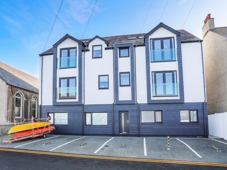A white and blue three story building with six large windows and five empty parking spaces with two kayaks on a trailer at Brig-Y-Don 4 in Rhosneigr