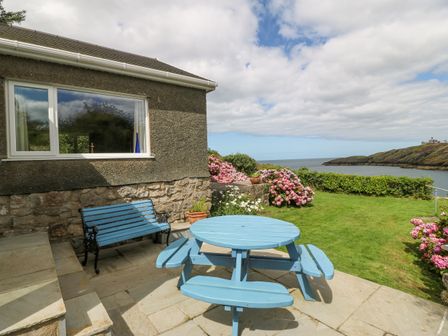 An outdoor patio with a blue wooden picnic table and a blue bench near a house with a stone wall overlooking a garden and a body of water at The Headland in Llaneilian near Amlwch