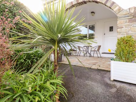 A porch area with a round table and two chairs next to a white door and window surrounded by green plants and a stone arch at EdyVean in Portmellon near Mevagissey