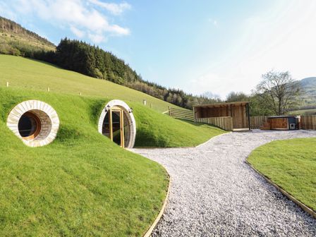 A hillside home with round windows and door next to a gravel path and wooden shelter at Nant y Garnedd Mallwyd near Dinas Mawddwy