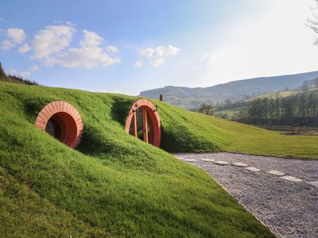 A grassy earth house with round doors and windows in a rural setting with hills and a gravel path at Nant yr Esgair Mallwyd near Dinas Mawddwy