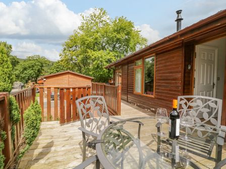 An outdoor wooden deck with a glass table and metal chairs with leaf designs a bottle of wine and two wine glasses and wooden buildings and trees in the background at Kirkstone Lodge Limefitt Park near Windermere