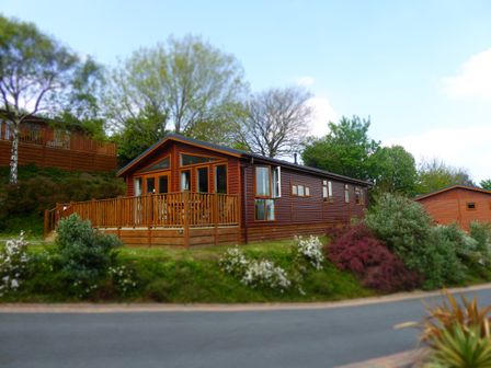 A wooden cabin with a fenced deck surrounded by bushes and trees at 23 Bossiney Bay in Tintagel