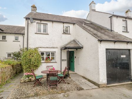 A white cottage exterior with a green door a round wooden table and chairs and a black garage door at Columbine Cottage in Hawkshead