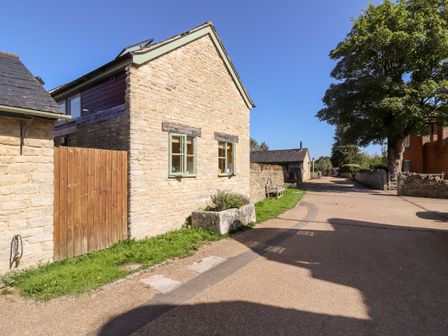 A stone house with two windows and a wooden gate next to a paved street with a bench and trees at Mill View in Cotswold Water Park