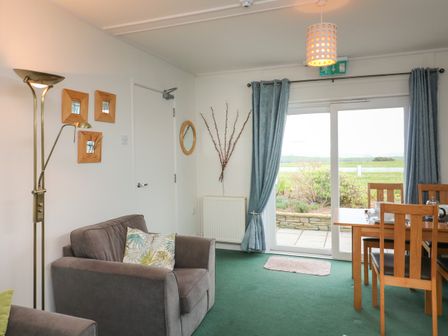 A living area with a gray armchair and wooden dining table near sliding glass doors with blue curtains at Fairway Lodge in Bigbury-On-Sea