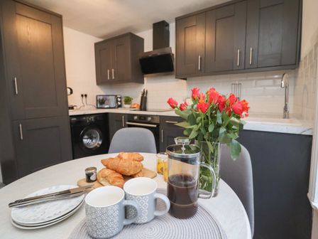 A kitchen with dark cabinets a washing machine an oven and a table set with croissants mugs a coffee press and red flowers at Monteva Whitby