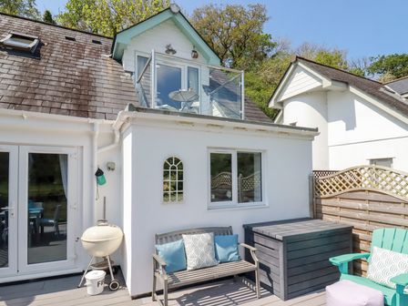 An outdoor patio area with a bench, cushions, a barbecue grill, a wooden storage box, a turquoise chair, and a small balcony above at Penpoll in Little Petherick Creek near Padstow