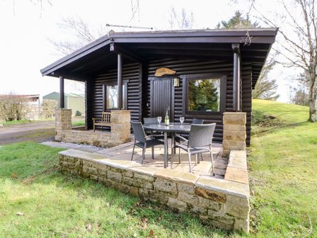 A dark wooden cabin with stone pillars and an outdoor patio table with chairs and wine glasses at Alwinton Lodge in Longframlington
