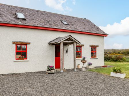 The exterior of a white house with red window frames and door surrounded by gravel and grass with flower pots at Cob Cottage in Kilmore County Wexford