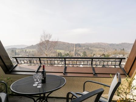 A balcony with a metal table holding a bottle and four wine glasses overlooking a town and hills at Rothay in Ambleside