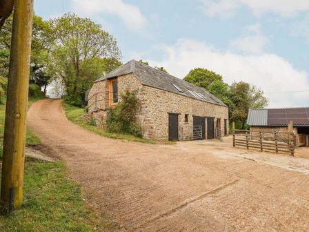 A stone barn with black doors and a driveway surrounded by trees at The Coach House at Thorn Farm in Bridford near Moretonhampstead