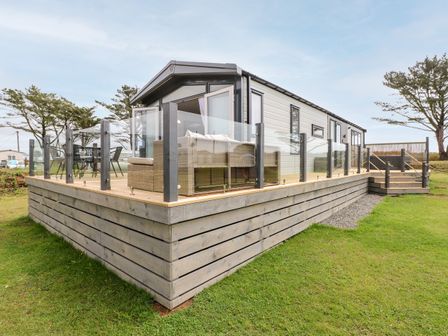 A raised wooden deck with glass railing around a modular home with outdoor furniture on the deck at Puffin Lodge in Broad Haven