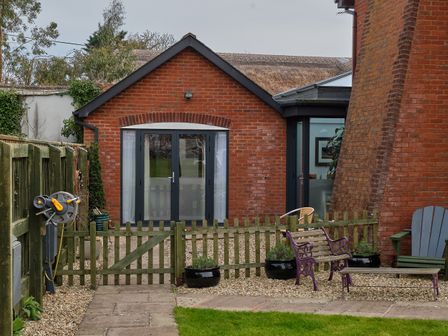 A backyard with a small gate, wooden bench, potted plants, hose reel, and a brick building with glass doors at Farriers Retreat Budleigh Salterton