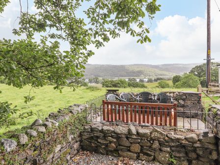 An outdoor seating area with plastic chairs and a wooden bench enclosed by a stone wall overlooking green fields and a lake at 1 Lake View in Coniston
