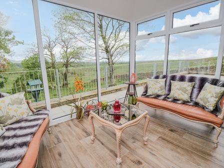 A sunroom with two sofas a glass table with drinks and a view of trees and fields at Torties Cottage in West Hall