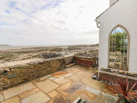 A stone patio with a low stone wall and decorative window on a white building overlooking a rocky beach at 1 Salt Cottage in Port Eynon