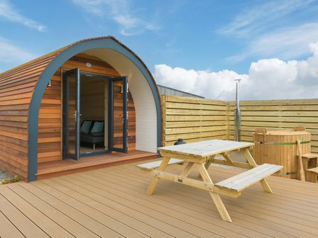 A wooden pod with open double doors a picnic table and a wooden hot tub on a deck at Green Fields in St Buryan