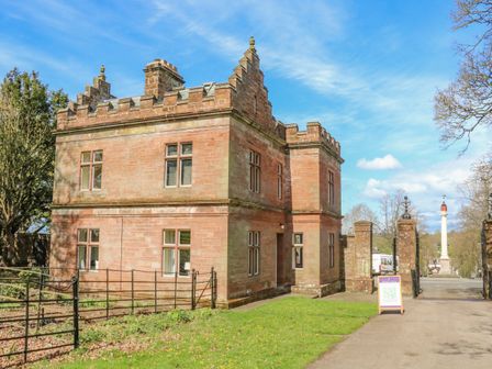 An old stone building with battlements near a gated entrance and a monument in the background at North Lodge in Appleby-In-Westmorland