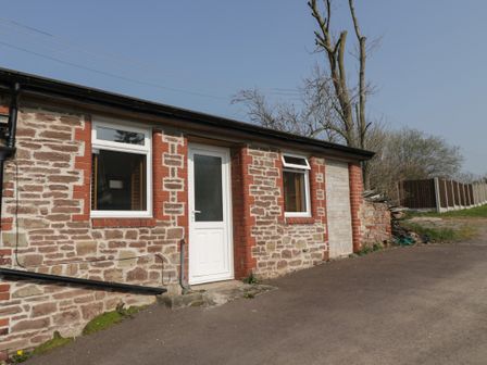 A single-story stone building with two windows and a white door near a paved area at Netherend Inn in Woolaston