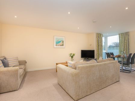 A living room with beige sofas a wooden coffee table a television and a dining table with chairs with curtains and a window at Golden Sands in Carbis Bay