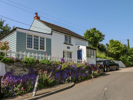 A house with a white picket fence and flowers along the stone wall next to a road with a black car parked nearby at Hillside in Crafthole