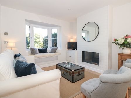 A living room with a white sofa blue cushions a gray armchair a wooden table with flowers a round mirror above a fireplace and a trunk coffee table at Marroy in Salcombe