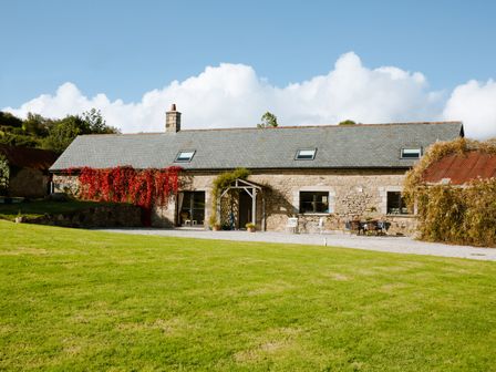A stone barn with a slate roof surrounded by grass and climbing plants at Higher Weddicott Barn in Chagford