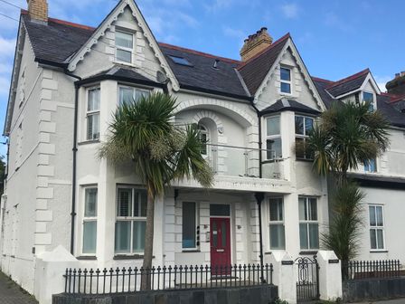 A white two story house with a red door two palm trees and a metal fence in front at Apartment 2, Gorwel Glas in Llanbedrog