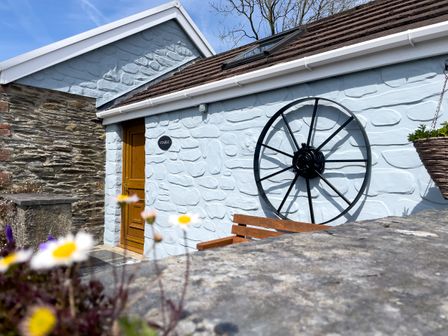 An outdoor view of a stone building with a wooden door a large black metal wheel mounted on the wall and flowers in the foreground at The Stable @ Canllefaes in Penparc near Cardigan