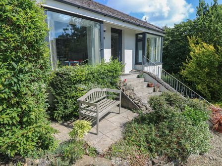 A garden bench on a concrete patio surrounded by bushes outside a house with steps and glass windows at Pinewood in Saundersfoot