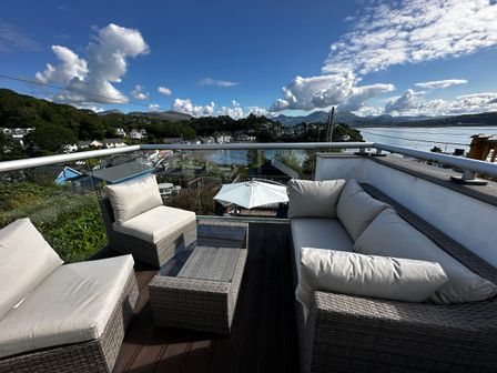 An outdoor patio with cushioned wicker furniture and a glass table overlooking a coastal town and mountains at Alltwen in Borth-y-Gest