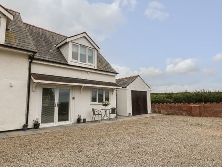A white house with two chairs and a table on a paved area next to a gravel driveway at Bryn Eglwys Bach in Penmynydd near Llanfairpwllgwyngyll