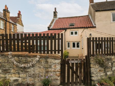A small house with a red tile roof behind a stone wall and wooden fence with a gate labeled Stonewall in Sandsend