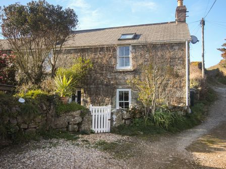 A stone cottage with a slate roof and white gate next to a gravel driveway and small garden at Rose Cottage in Lower Bostraze near St Just