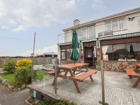 An outdoor seating area with wooden picnic tables and closed umbrellas outside a building with stone facade and glass railing at Morlyn Guest House Apartment in Llandanwg near Harlech