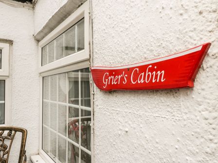 A red wooden sign reading Griers Cabin mounted on a white exterior wall with windows and a metal chair nearby at Griers Cabin in Whitby