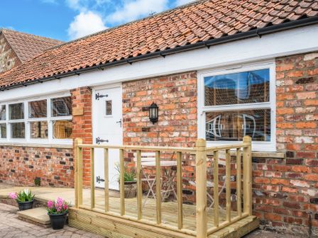 A brick house with a small wooden porch and white door and windows with flower pots outside at The Old Tractor Shed in Skipsea